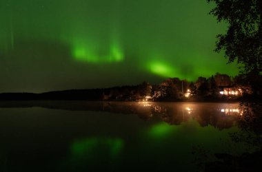 İsveç 'in Stockholm yakınlarındaki sakin bir gölün üzerindeki kuzey ışıkları suya yansıdı. Evler gölü de sarar.