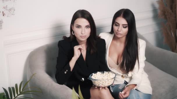 Two friends stylish brunette women are sitting on a sofa and eating popcorn — Stock Video