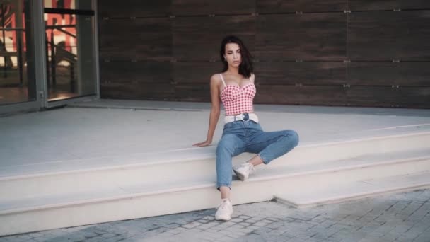 Молода тонка біла брюнетка модель жінки в повсякденному одязі сидить на сходах — стокове відео