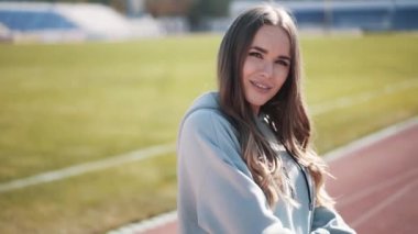 Ağır çekimde, gri eşofmanlı, beyaz bir kız stadyumda poz veriyor.