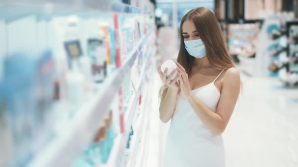Милая белая красивая девушка в медицинской маске выбирает крем для рук — стоковое видео