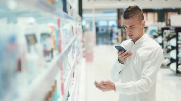 Guy fotografía un frasco con cosméticos en un smartphone en un supermercado — Vídeos de Stock