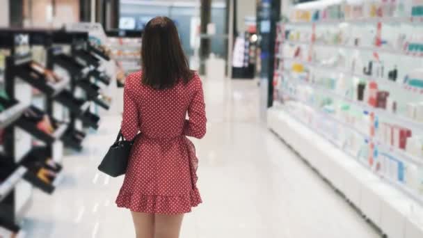 Жінка у червоній сукні ходить по полицях у косметичному бутіку. — стокове відео