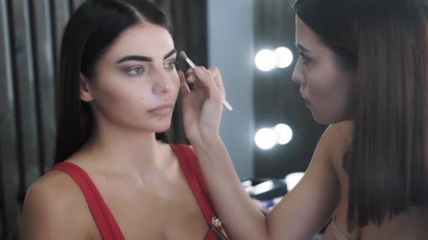 Гарненька жінка візажистка застосовує макіяж до молодої красивої брюнетки — стокове відео
