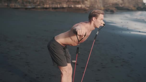 Молодой кавказский спортсмен делает упражнения с эластичной лентой на берегу моря. — стоковое видео