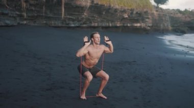 Deniz kıyısında direniş bandıyla egzersiz yapan genç ve güçlü bir adam..