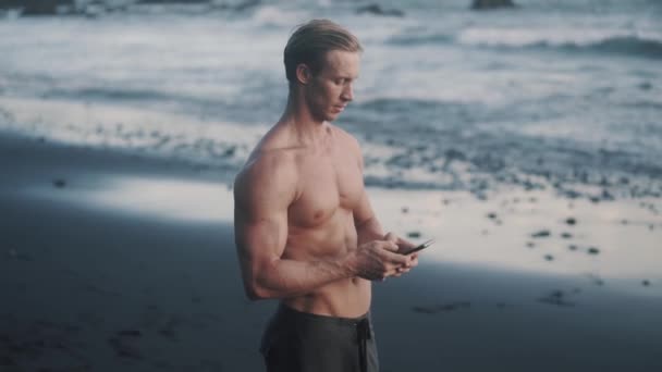 Topless young sportsman uses his smartphone at the sea shore. — Stock Video