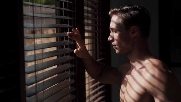 Młody biały mężczyzna spogląda przez okno przez żaluzje na słońce. — Wideo stockowe