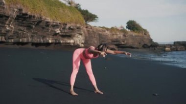 Spor giyimli genç beyaz bir kadın deniz kıyısında yarım yamalak yamaç yapıyor..