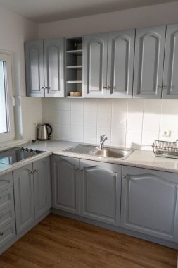 Gri dolapları, seramik tenceresi ve paslanmaz çelik lavabosu olan modern bir mutfak. Pencereden gelen güneş ışığıyla aydınlatılıyor. Minimalist tasarım ve döşeme döşemeleriyle rahat ve işlevsel ev tasarımı, gayrimenkul teması için uygun, int