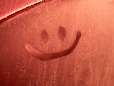 Kırmızı renkli sisli ya da soğuk bir yüzeye çizilmiş neşeli bir gülen yüz, sıcak ışıklandırmayı dokulu bir arka planla birleştirerek neşeli, canlandırıcı ve pozitif bir görsel ifade yaratır..