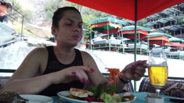 Bir kız, nehir kıyısındaki küçük bir şelalede restoranda yemek yiyor.