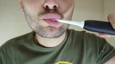 Sakallı bir adam, dişlerini aynada ağzı açık fırçalar. Elektrikli diş fırçasında diş macunu.