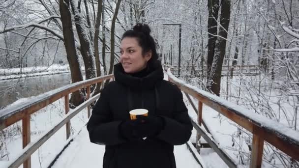 Vacker ung flicka går på vintern i parken med ett glas kaffe i händerna — Stockvideo