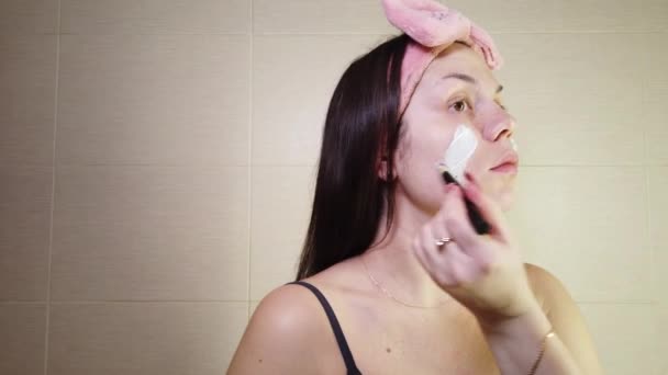 化粧ブラシを使用して顔にアルギン酸マスクを適用ピンクのヘッドバンドと若いブルネット 自宅でのスキンケア手順 — ストック動画