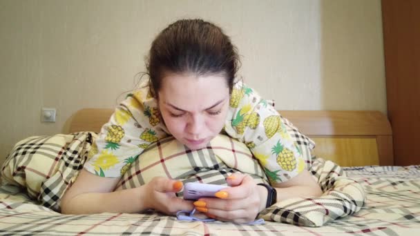優しい女性がベッドに横になってスマートフォンにメッセージを送る — ストック動画