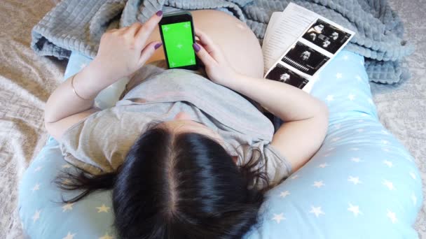 En gravid kvinna ligger i sängen med en telefon i handen som håller hennes mage, täcker hennes ben med filt, ovanifrån — Stockvideo