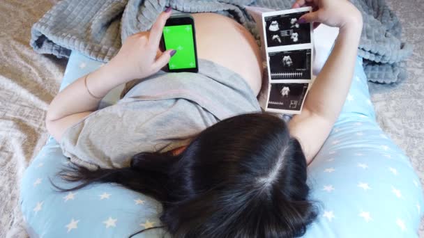 妊娠中の女性は 毛布で彼女の足をカバーし 彼女の胃を保持彼女の手の電話でベッドに横たわっているトップビュー — ストック動画