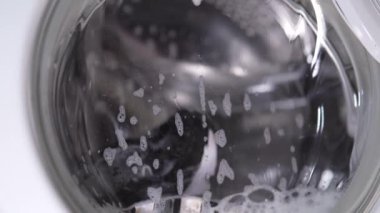 Beyaz çamaşır makinesi kirli ayakkabıları yıkar. Ev yapımı çamaşır makinesinde ayakkabı yıkamak. Cam kapıda çamaşır tozu köpüğü var..