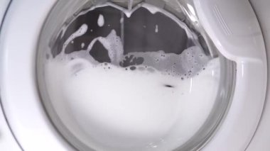 Beyaz çamaşır makinesi kirli ayakkabıları yıkar. Ev yapımı çamaşır makinesinde ayakkabı yıkamak. Cam kapıda çamaşır tozu köpüğü var..