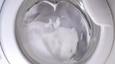 Beyaz çamaşır makinesi kirli ayakkabıları yıkar. Ev yapımı çamaşır makinesinde ayakkabı yıkamak. Cam kapıda çamaşır tozu köpüğü var..