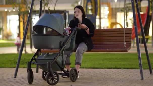 Mom with a stroller is sitting on a swing in the courtyard of a residential building — Stock Video