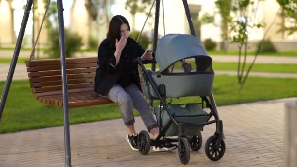 Moeder met een wandelwagen zit op een schommel op de binnenplaats van een woongebouw — Stockvideo