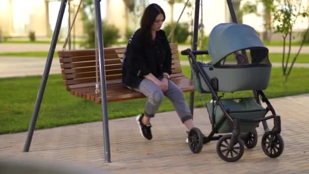 Мама с коляской сидит на качелях во дворе жилого дома — стоковое видео