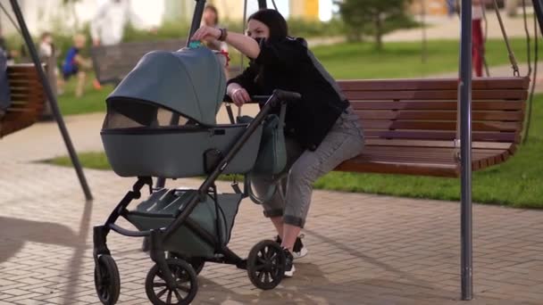 Moeder met een wandelwagen zit op een schommel op de binnenplaats van een woongebouw — Stockvideo