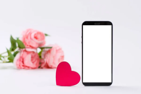 Pembe kalp ve beyaz arka planda beyaz ekranlı akıllı telefon. Arka planda bir buket pembe gül var. Mesajlarla ilan edilen aşk kavramı