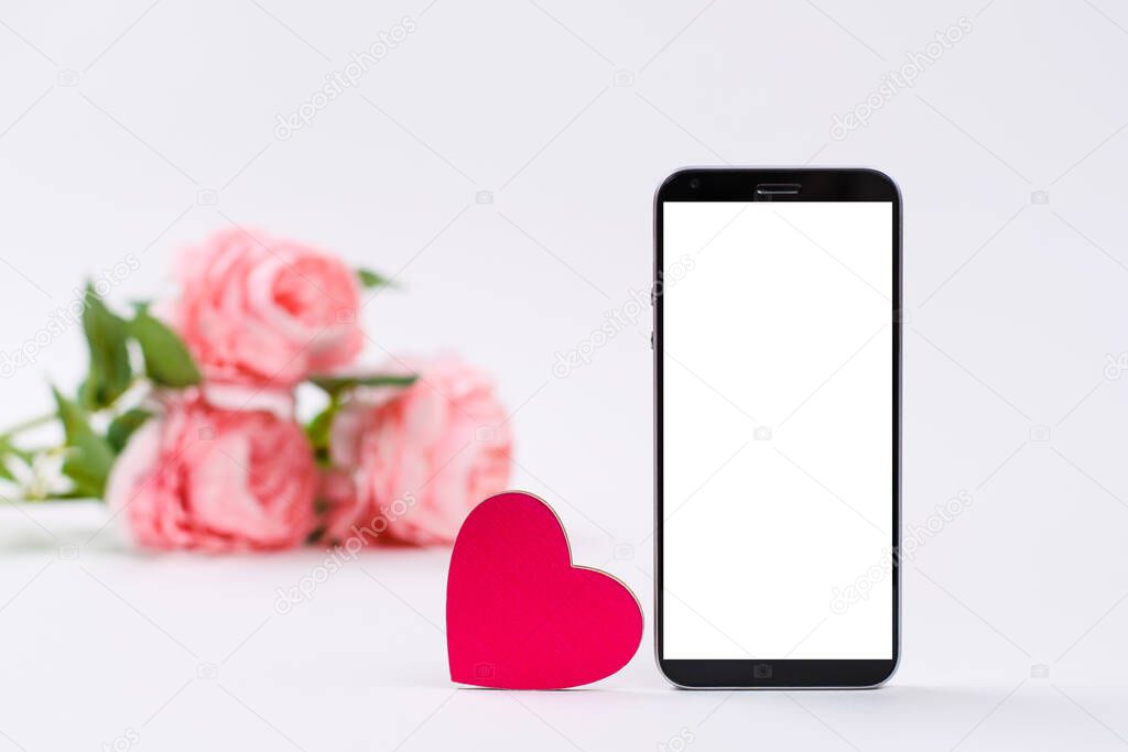 Pink heart and smartphone with blank white display against white background. in the background a bouquet of pink roses. Concept of declaration of love through messages