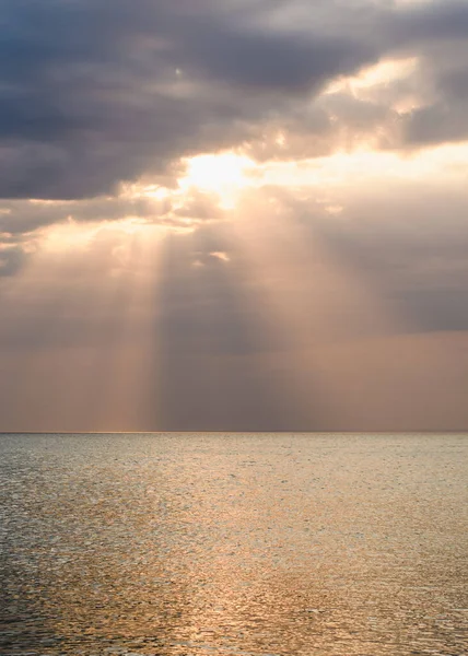 Denizin üzerinde gün batımı. Güneş ışınları bulutların arkasından parlıyor. Dikey çerçeve.