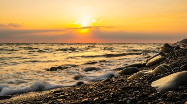 Denizde gün batımı. Çakıl taşı plajı ve gün batımında sörf. Uzun süre ateş edildi. Suyun kenarına ve çakıl taşlarına odaklan..