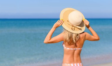 Hasır şapkalı ince bir kız güneşli bir günde deniz kıyısında duruyor. Tropikal iklimde plaj tatili.