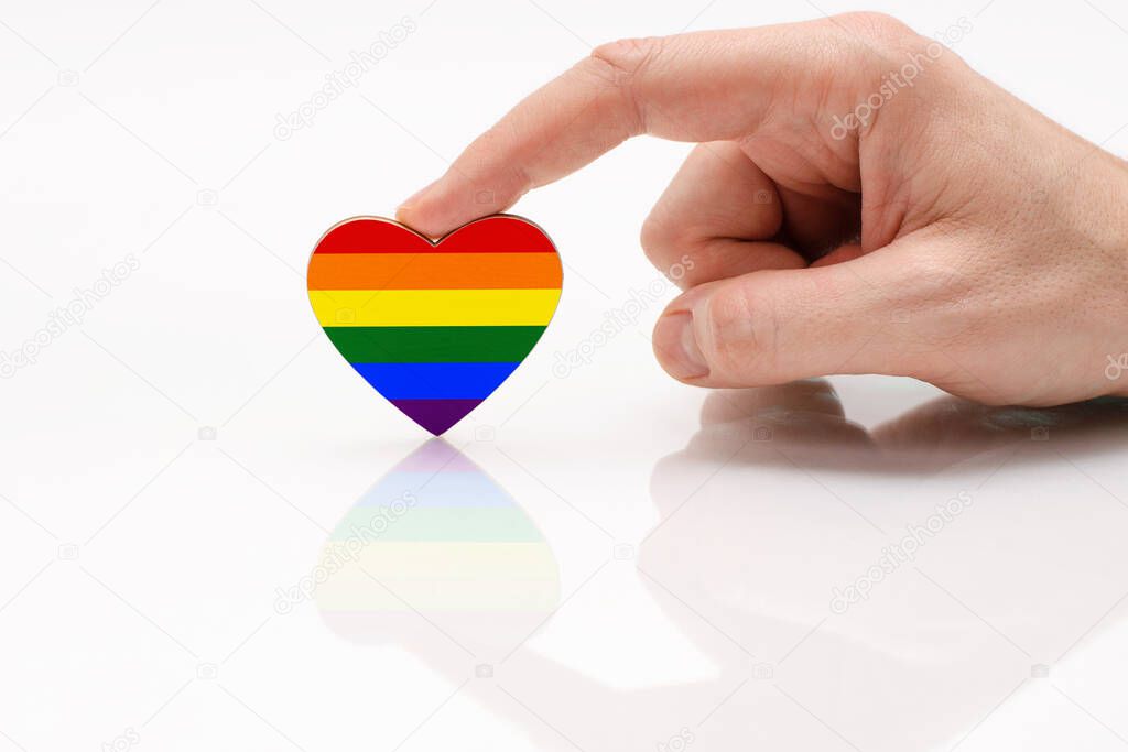 Gay Pride Flag in the shape of a heart. A man's hand holds a heart in the shape of the LGBT flag on a white glass surface. The concept of tolerance and equality for LGBT people
