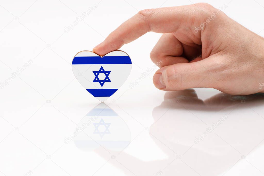 Love and respect the Israel. A man's hand holds a heart shaped like an Israel flag on a glass white surface. The concept of Israeli patriotism and pride.