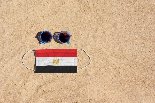 A medical mask in the color of the Egypt flag lies on the sandy beach next to the glasses. The concept of organizing a safe beach holiday in Egypt during the coronavirus pandemic.