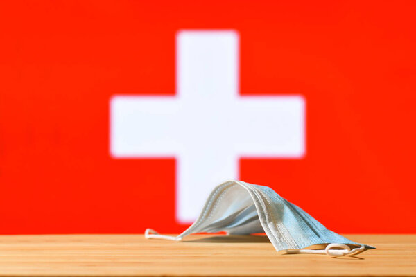 A medical mask lies on the table against the background of the flag of Switzerland. The concept of a mandatory mask regime for residents of the country and tourists in Switzerland during a pandemic.