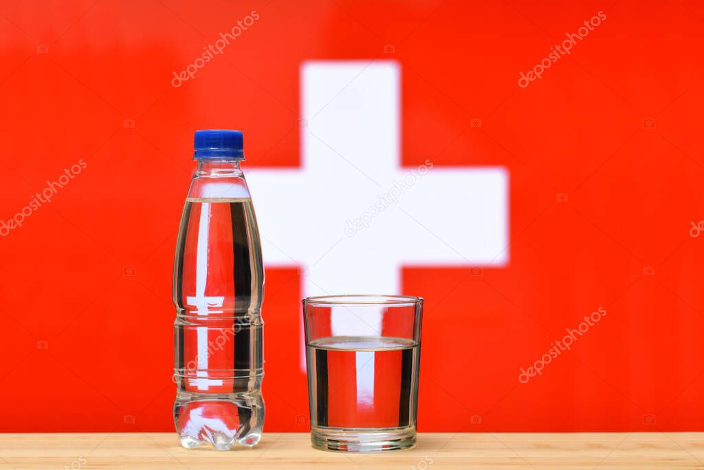 A bottle of clean drinking water and a glass stand on the table against the background of the flag of Switzerland. A concept for the supply of clean drinking water in Switzerland.