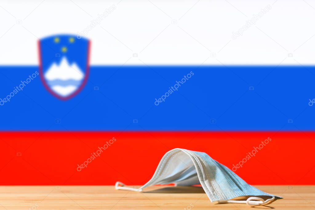 A medical mask lies on the table against the background of the flag of Slovenia. The concept of a mandatory mask regime for residents of the country and tourists in Slovenia during a pandemic.
