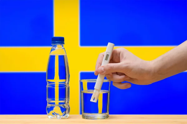 水テスター付きの手は スウェーデンの国旗を背景に澄んだ水のガラスで測定を行います スウェーデンにおける飲料水供給のテストと評価 — ストック写真