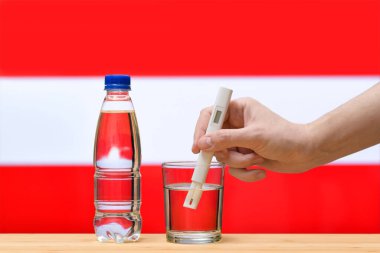 Su testi yapan bir el, Avusturya bayrağının arka planına karşı bir bardak temiz suda ölçüm yapar. Avusturya 'daki içme suyu kaynaklarının test ve değerlendirmesi.