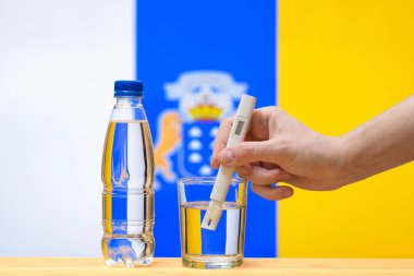 Su testi yapan bir el, bir bardak temiz suda Kanarya Adaları bayrağının arka planına karşı bir ölçüm yapar. Kanarya Adaları 'ndaki içme suyu kaynaklarının test ve değerlendirmesi.