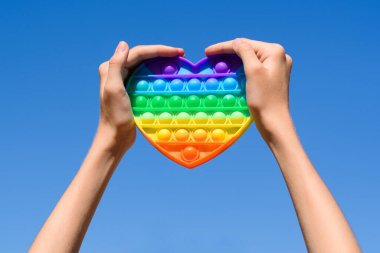 Toy pop it in the shape of a heart on a background of the sky. Heart in the colors of the rainbow in the hands.