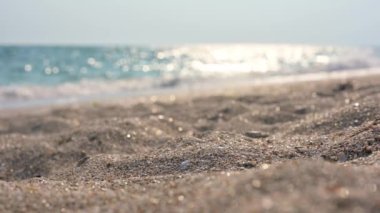 Kumsal ve deniz dalgaları güneşli bir günde. Ön plandaki kuma yakın çekim ve seçici odaklanma. Deniz dalgaları odak dışı, röfleli ve bokeh.