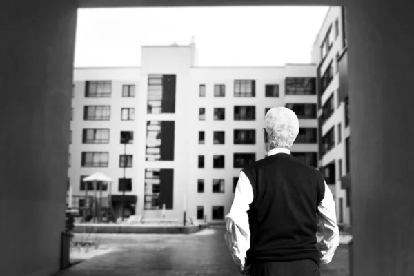 An elderly man looks at a construction site, at home. The view from the back. Black and white.
