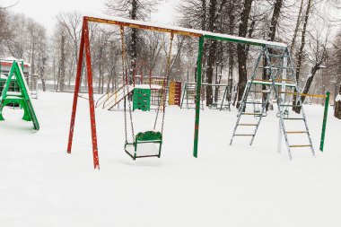 Kar yağışında terk edilmiş çocuk parkı salıncakları, renkli ekipmanların ve beyaz kış manzarasının kontrastını vurguluyor.