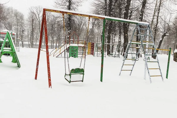Kar yağışında terk edilmiş çocuk parkı salıncakları, renkli ekipmanların ve beyaz kış manzarasının kontrastını vurguluyor.