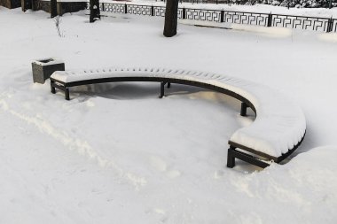 Sessiz bir kış parkında, sakin kentsel tasarım ve soğuk mevsimsel atmosfer gösteren, taze karla kaplı kıvrımlı ahşap bir bank..