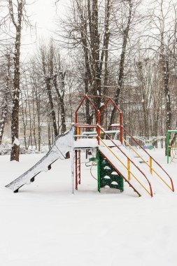 Terk edilmiş slayt seti bir oyun parkında kar altında gömülü, kış sükunetini ve açık hava oyunlarına sezonluk molası simgeliyor..